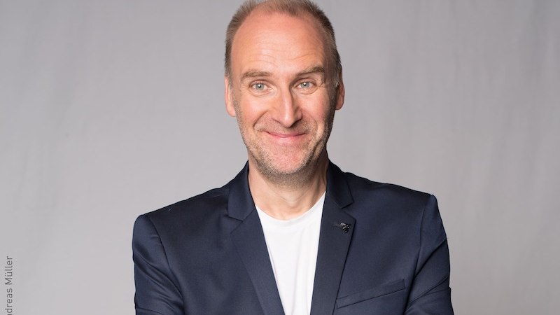 Ein Mann in einem dunklen Anzug mit wei&szlig;em T-Shirt l&auml;chelt vor einem grauen Hintergrund., &copy; Verein der Freunde des Lions Club Kornwestheim e.V.