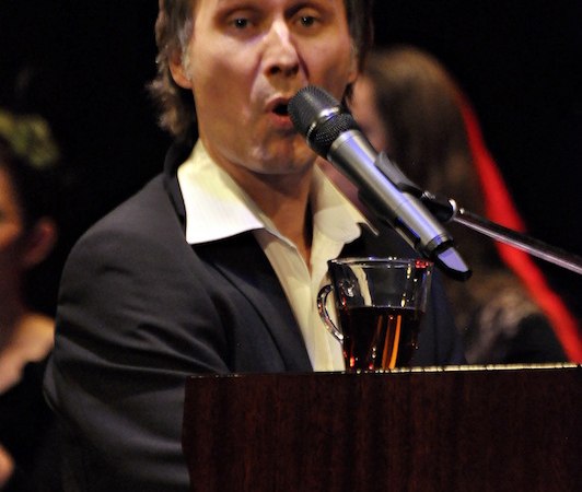 Ein Mann in Anzug singt am Klavier in ein Mikrofon. Im Hintergrund sind unscharfe Personen zu sehen., &copy; Touristik Bad Wildbad GmbH