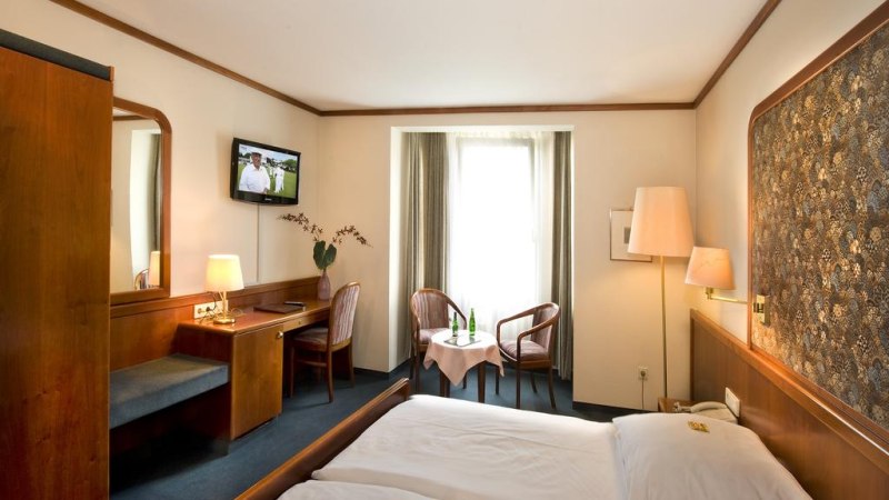 Gem&uuml;tliches Hotelzimmer mit Doppelbett, Fernseher, Schreibtisch und Sitzbereich vor einem Fenster. Holzm&ouml;bel und warme Beleuchtung dominieren den Raum., &copy; Hotel am Schelztor
