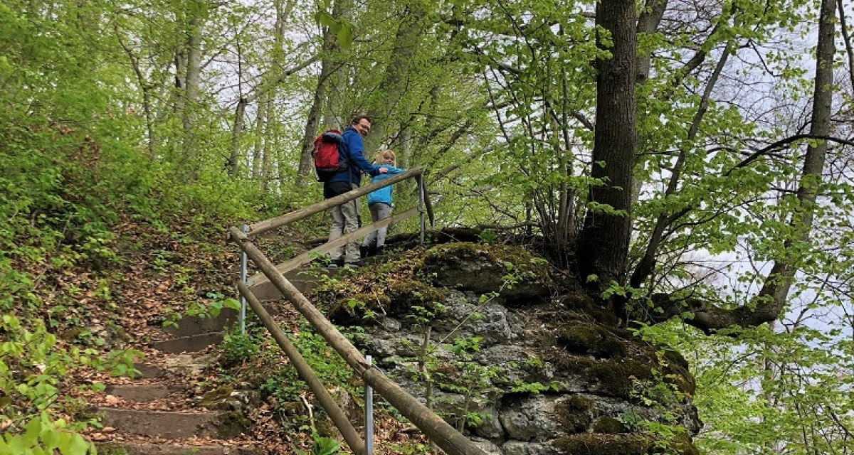 Zwei Wanderer stehen auf einem bewaldeten Pfad mit Geländer. Der Weg führt durch einen grünen Wald mit Laub und Felsen., © Touristik und Marketing GmbH