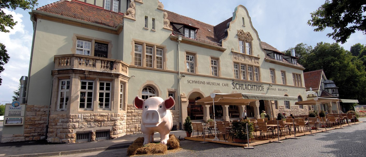 Historisches Gebäude mit Schweinemuseum und Café Schlachthof. Große Schweinefigur steht davor, umgeben von Tischen und Stühlen im Außenbereich., © Buschwerk Mediendesign-Wilhelmer Gastronomie