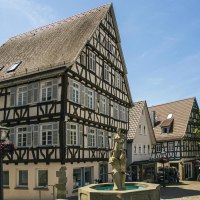 Fachwerkh&auml;user am Marktplatz von Backnang, mit einem Brunnen im Vordergrund und B&auml;umen im Hintergrund. Menschen spazieren und sitzen in Caf&eacute;s., &copy; Stuttgart-Marketing GmbH, Sarah Schmid