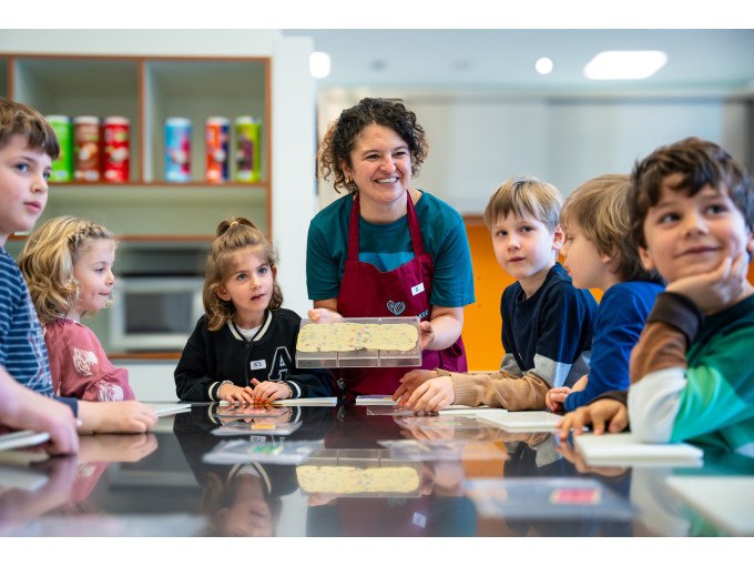Eine Frau in Sch&uuml;rze zeigt Kindern eine Schokoladentafel. Die Kinder sitzen um einen Tisch in einer modernen K&uuml;che., &copy; Alfred Ritter GmbH &amp; Co. KG | Waldenbuch