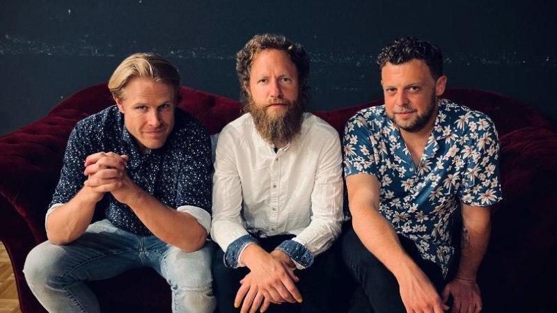 Drei Männer sitzen auf einem roten Sofa, lässig gekleidet, vor einem dunklen Hintergrund. Sie tragen Hemden mit Mustern und Jeans., © Foto Daniel Mudrack Drei Männer sitzen auf einem roten Sofa, lässig gekleidet, vor einem dunklen Hintergrund. Sie tragen Hemden mit Mustern und Jeans., © Foto Daniel Mudrack