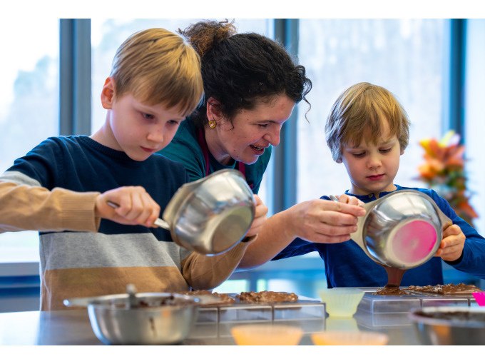 Zwei Kinder und eine Frau gießen Schokolade in Formen. Sie sind in einer Küche mit großen Fenstern., © Alfred Ritter GmbH & Co. KG | Waldenbuch Zwei Kinder und eine Frau gießen Schokolade in Formen. Sie sind in einer Küche mit großen Fenstern., © Alfred Ritter GmbH & Co. KG | Waldenbuch