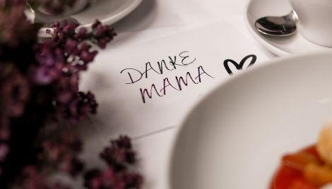 Ein liebevoll gedeckter Tisch mit einem Zettel, auf dem 'Danke Mama' steht. Blumen und Geschirr sind im Hintergrund zu sehen., &copy; Muttertatgs.Brunch. Foto: Teamwerk