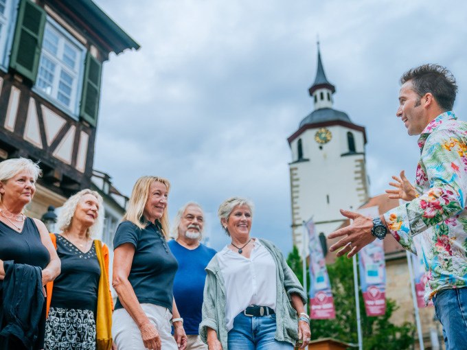 Eine Gruppe von Menschen lauscht einem Stadtf&uuml;hrer vor einem Fachwerkhaus und einem Turm mit Uhr. Der Himmel ist bew&ouml;lkt., &copy; Stadt Waldenbuch