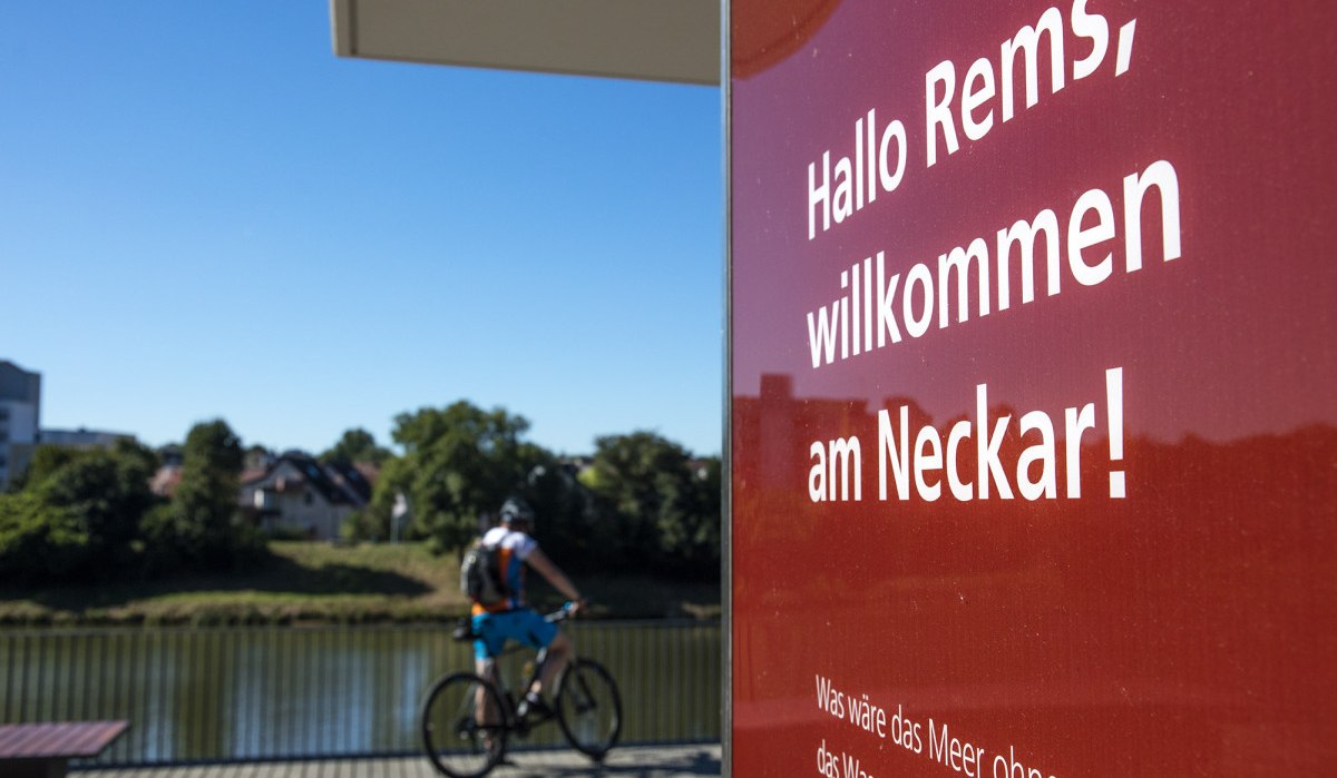 Schild mit 'Hallo Rems, willkommen am Neckar!' im Vordergrund, Radfahrer am Flussufer im Hintergrund. Schild mit 'Hallo Rems, willkommen am Neckar!' im Vordergrund, Radfahrer am Flussufer im Hintergrund.