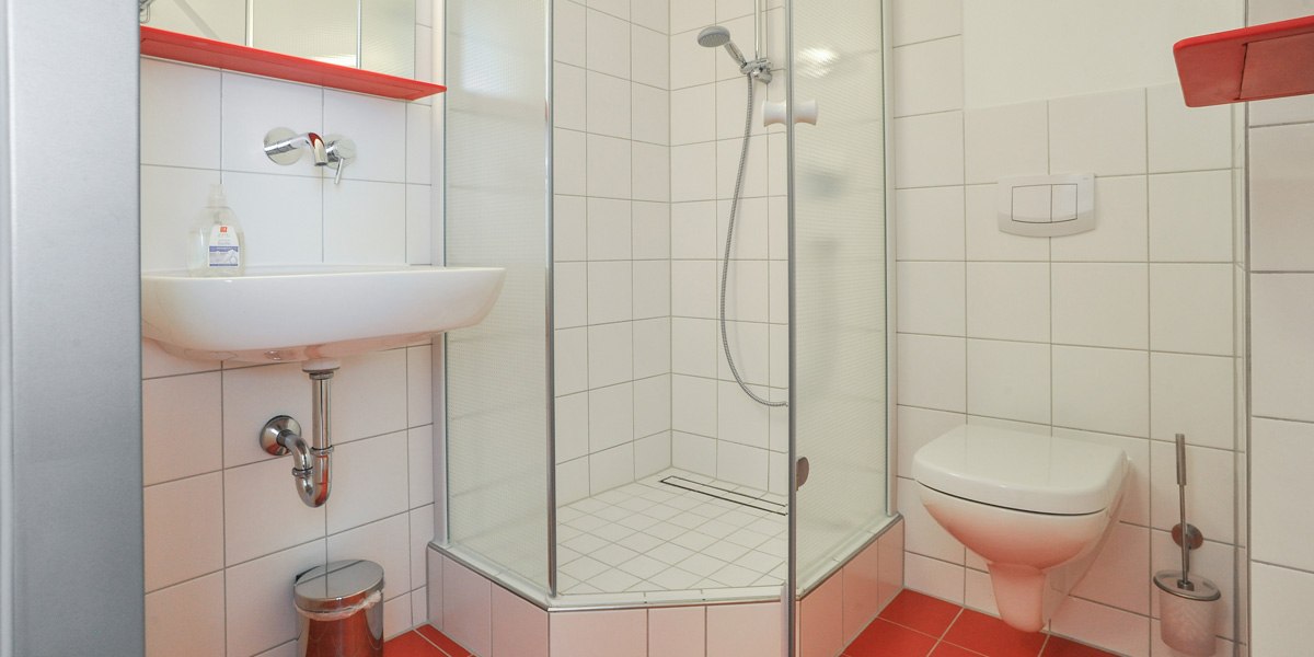 Ein modernes Badezimmer mit weiß gefliesten Wänden, einer Dusche, einem Waschbecken und einer Toilette. Rote Akzente und ein Mülleimer sind sichtbar., © Gästehaus ABZ Stuttgart Ein modernes Badezimmer mit weiß gefliesten Wänden, einer Dusche, einem Waschbecken und einer Toilette. Rote Akzente und ein Mülleimer sind sichtbar., © Gästehaus ABZ Stuttgart