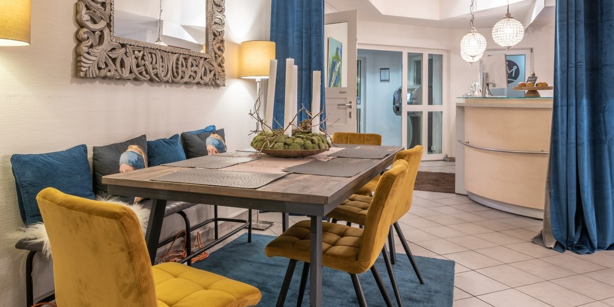 Gemütliche Lounge mit Esstisch, gelben Stühlen, blauem Teppich und dekorativem Spiegel. Im Hintergrund eine Empfangstheke und moderne Beleuchtung., © Hotel Princess