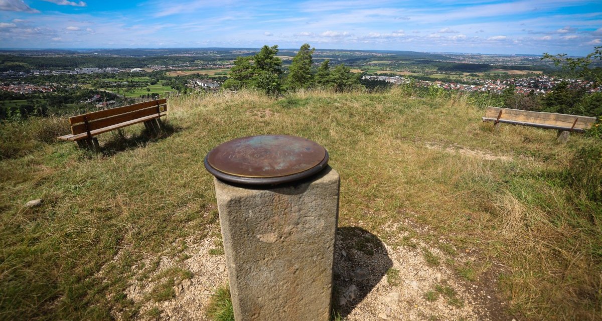 Aussichtspunkt mit zwei Bänken und einer Panoramatafel auf einem Hügel. Im Hintergrund weite Landschaft und blauer Himmel. Aussichtspunkt mit zwei Bänken und einer Panoramatafel auf einem Hügel. Im Hintergrund weite Landschaft und blauer Himmel.