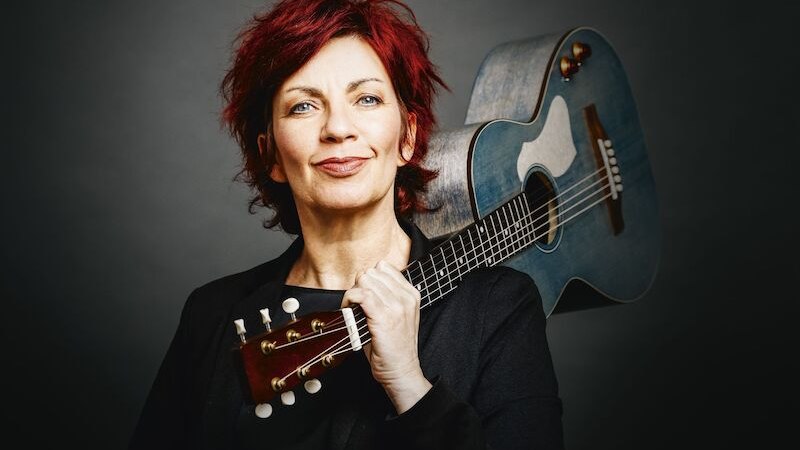 Frau mit rotem Haar hält eine Gitarre über der Schulter und lächelt. Dunkler Hintergrund., © Mauerwerk GmbH Frau mit rotem Haar hält eine Gitarre über der Schulter und lächelt. Dunkler Hintergrund., © Mauerwerk GmbH