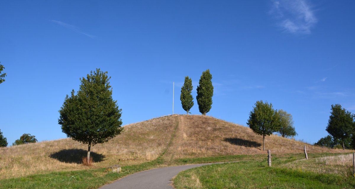Ein Hügel mit einem Pfad führt zu drei Bäumen und einem Fahnenmast. Der Himmel ist klar und blau., © Remstal Tourismus e.V. Ein Hügel mit einem Pfad führt zu drei Bäumen und einem Fahnenmast. Der Himmel ist klar und blau., © Remstal Tourismus e.V.