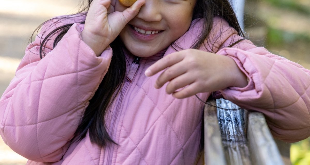 Ein Kind in rosa Jacke hält eine kleine Kugel mit Gesicht vor das Auge und lächelt fröhlich. Im Hintergrund ist ein Weg zu sehen., © Thomas Zehnder Ein Kind in rosa Jacke hält eine kleine Kugel mit Gesicht vor das Auge und lächelt fröhlich. Im Hintergrund ist ein Weg zu sehen., © Thomas Zehnder