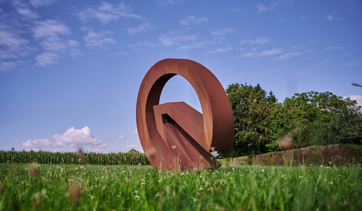 Eine große, rostfarbene Metallskulptur in Form eines Kreises steht auf einer grünen Wiese unter einem blauen Himmel., © Natur.Nah. Schönbuch & Heckengäu Eine große, rostfarbene Metallskulptur in Form eines Kreises steht auf einer grünen Wiese unter einem blauen Himmel., © Natur.Nah. Schönbuch & Heckengäu