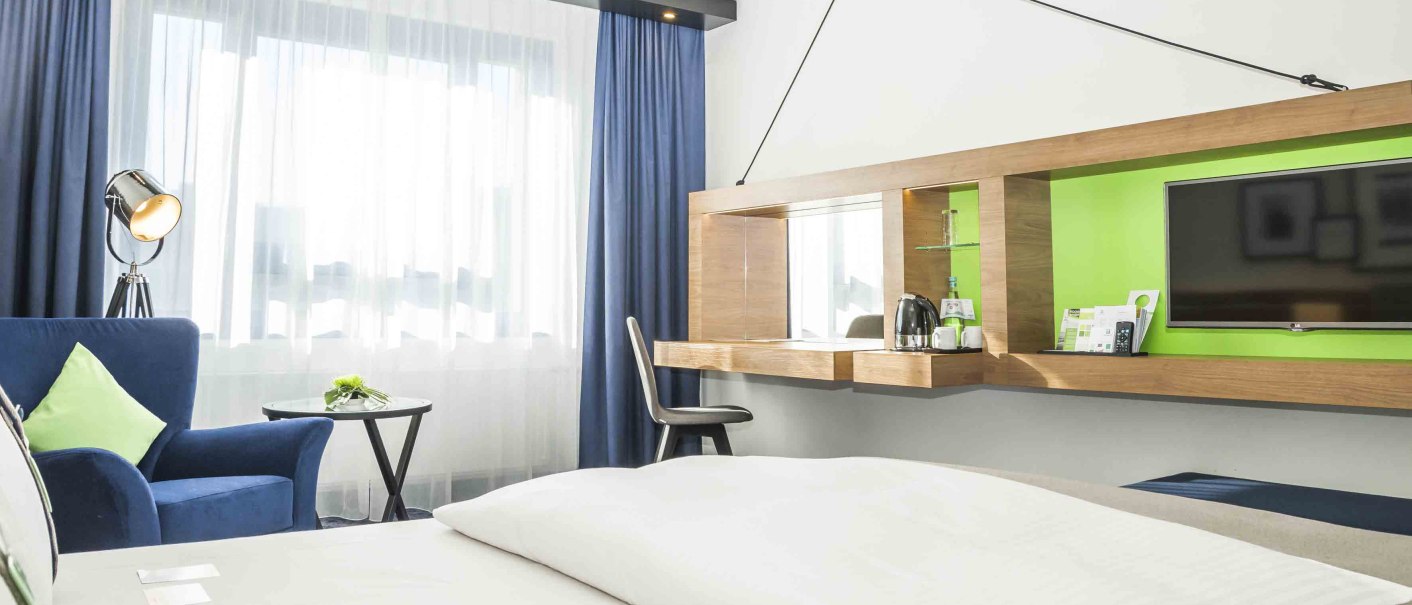 Modernes Hotelzimmer mit weißem Bett, blauem Sessel, Schreibtisch, Fernseher und großer Fensterfront mit blauen Vorhängen., © Chandler Photography Modernes Hotelzimmer mit weißem Bett, blauem Sessel, Schreibtisch, Fernseher und großer Fensterfront mit blauen Vorhängen., © Chandler Photography