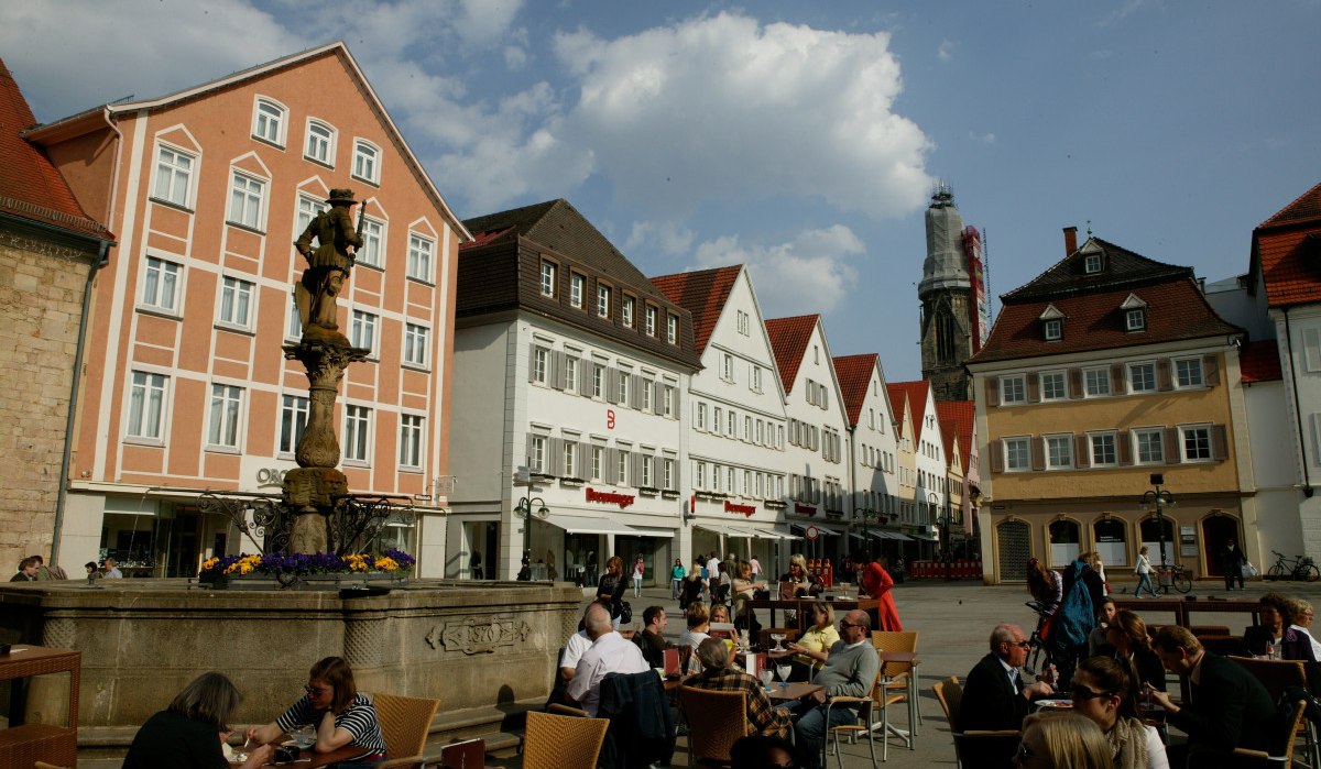 Der Reutlinger Marktplatz mit Menschen, die an Tischen sitzen. Historische Gebäude und ein Brunnen sind im Hintergrund zu sehen., © Sphäre Verlag