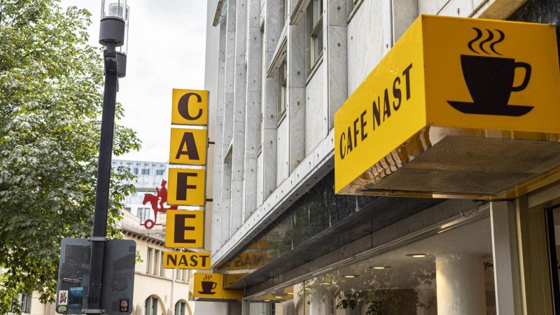 Gelbes Schild des Cafe Nast mit Kaffeetasse-Logo an einem Gebäude, umgeben von Bäumen., © SMG, Sarah Schmid