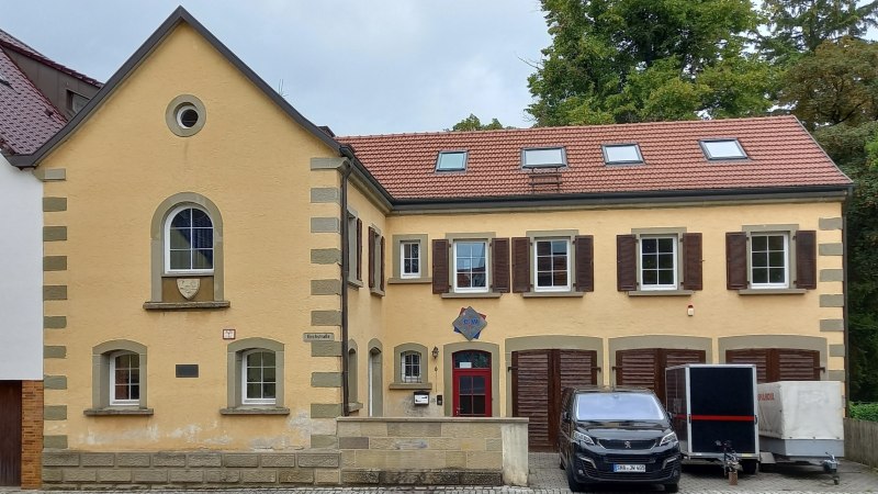 zweist&ouml;ckiges historisches Geb&auml;ude mit gelber Fassade und roten Dachziegeln. Im Untergescho&szlig; auf der rechten Seite drei rotbraune Doppelt&uuml;ren. Vor den T&uuml;ren stehen ein schwarzer PKW und zweiAnh&auml;nger, &copy; Petra Natzkowski