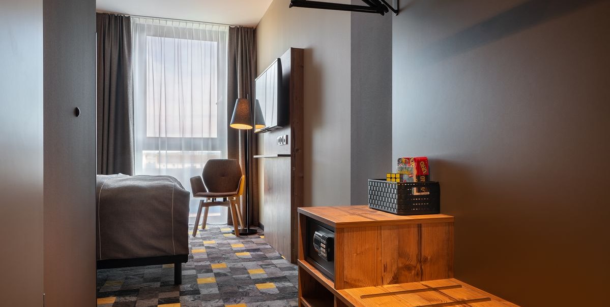 Modernes Hotelzimmer mit grauem Teppich, Bett, Sessel, Fernseher und Holzmöbeln. Ein Fenster mit Vorhängen lässt Tageslicht herein., © Novum Hospitality Modernes Hotelzimmer mit grauem Teppich, Bett, Sessel, Fernseher und Holzmöbeln. Ein Fenster mit Vorhängen lässt Tageslicht herein., © Novum Hospitality