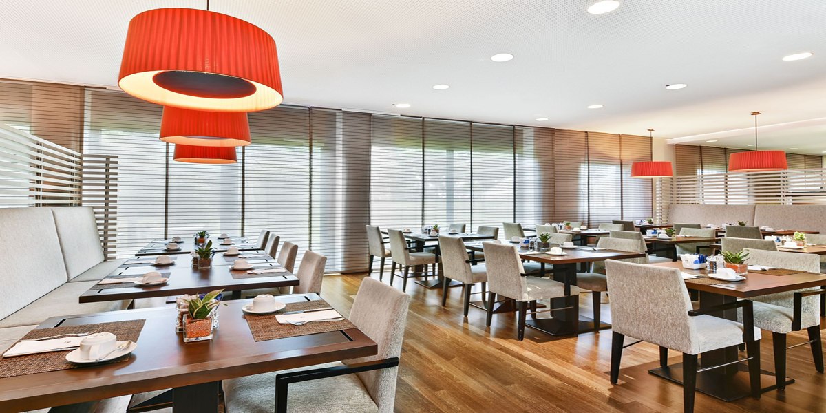 Ein moderner Frühstücksraum mit gedeckten Tischen, roten Hängelampen und großen Fenstern mit Jalousien., © NH Ludwigsburg
