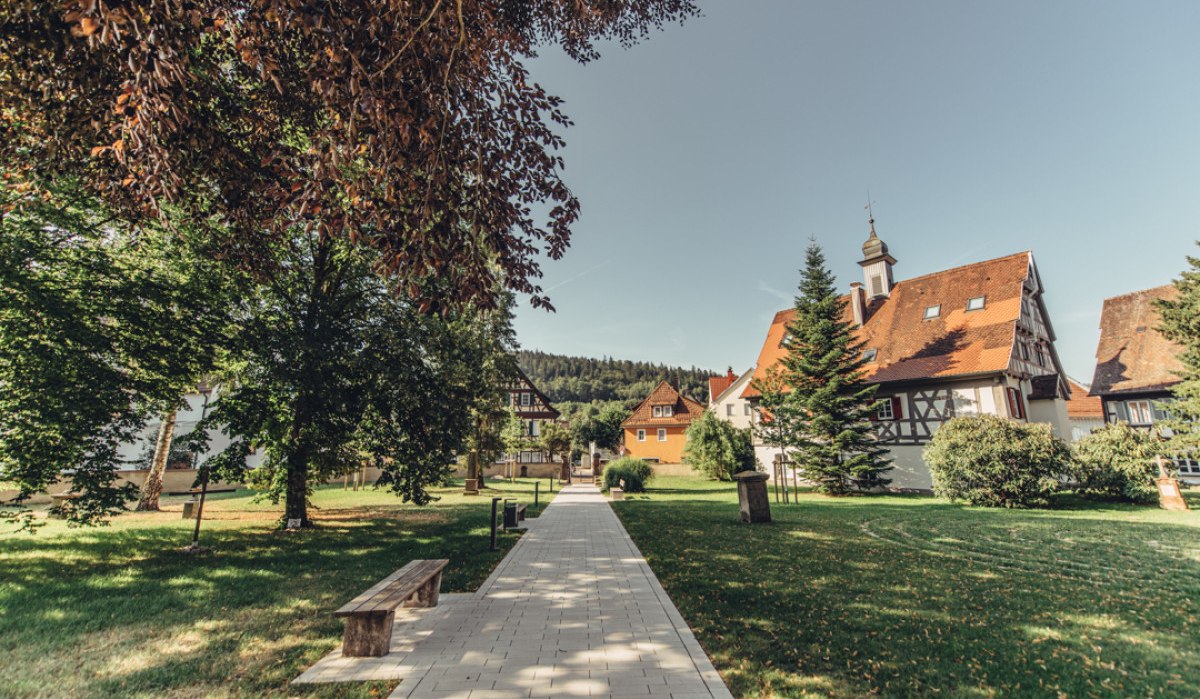 Gepflasterter Weg durch grünen Garten mit Bäumen und Bänken, umgeben von Fachwerkhäusern und Kirchturm im Hintergrund., © Tourismus Ostalb