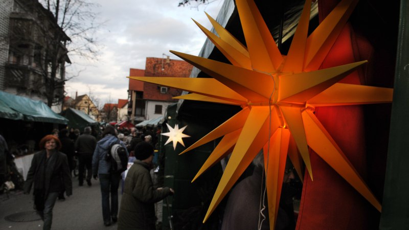 Ein Weihnachtsmarkt mit leuchtendem gelben Stern im Vordergrund. Menschen schlendern durch eine Gasse mit Ständen und Dekorationen., © Gemeinde Bad Boll