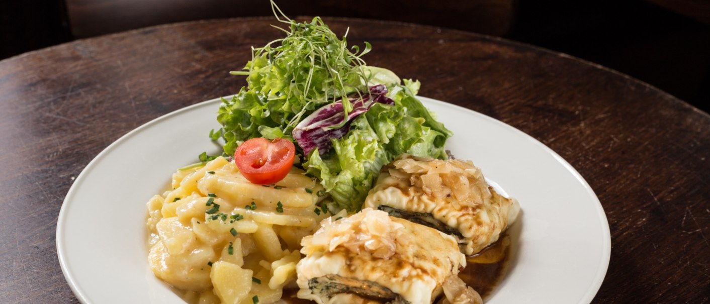 Ein Teller mit Maultaschen, Kartoffelsalat und grünem Salat, garniert mit einer Tomatenscheibe, auf einem Holztisch., © Amadeus, Stuttgart