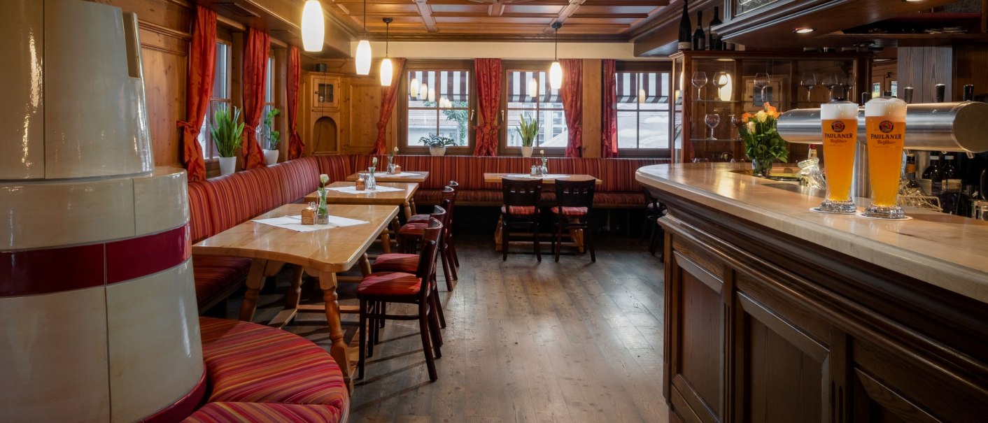 Gemütlicher Gastraum mit Holzmöbeln, roten Vorhängen und einer Bar. Zwei Biergläser stehen auf der Theke. Pflanzen dekorieren den Raum., © Hetzel Hotel Stuttgart Gemütlicher Gastraum mit Holzmöbeln, roten Vorhängen und einer Bar. Zwei Biergläser stehen auf der Theke. Pflanzen dekorieren den Raum., © Hetzel Hotel Stuttgart