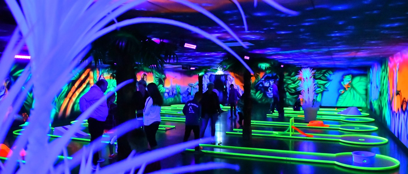 Neonbeleuchtete Minigolfanlage mit Dschungelthema. Menschen spielen auf leuchtenden Bahnen, umgeben von bunten Wandmalereien und Pflanzen., © Schwaben Park