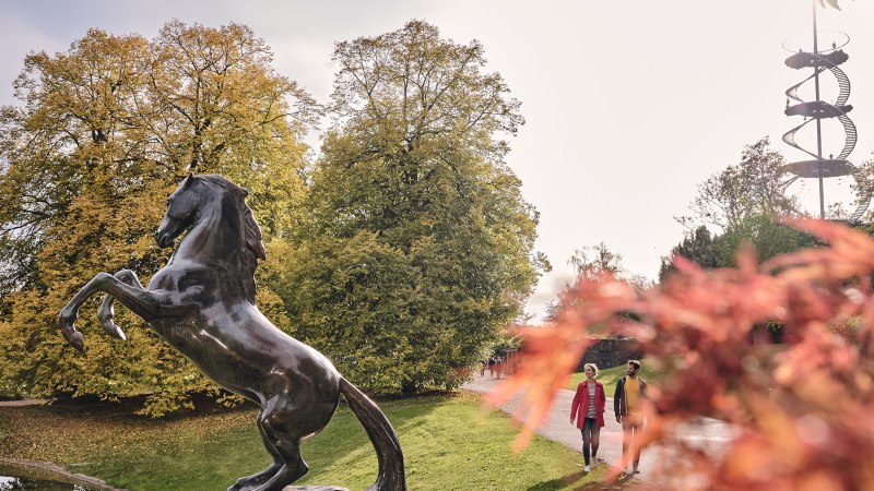 Bronzestatue eines aufsteigenden Pferdes im Höhenpark Killesberg, umgeben von herbstlichen Bäumen und einem spiralförmigen Aussichtsturm im Hintergrund., © SMG, Christoph Düpper Bronzestatue eines aufsteigenden Pferdes im Höhenpark Killesberg, umgeben von herbstlichen Bäumen und einem spiralförmigen Aussichtsturm im Hintergrund., © SMG, Christoph Düpper