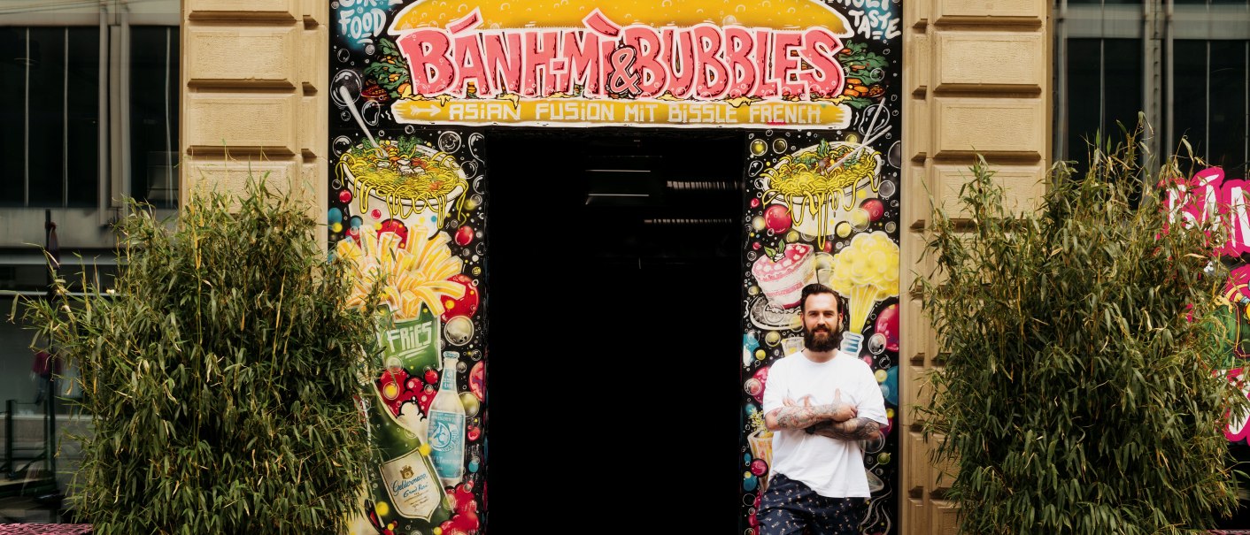 Ein Mann steht vor dem Eingang von 'Banh Mi & Bubbles', umgeben von bunten Graffiti und Pflanzen., © Banh Mi & Bubbles Restaurant / Bar Ein Mann steht vor dem Eingang von 'Banh Mi & Bubbles', umgeben von bunten Graffiti und Pflanzen., © Banh Mi & Bubbles Restaurant / Bar