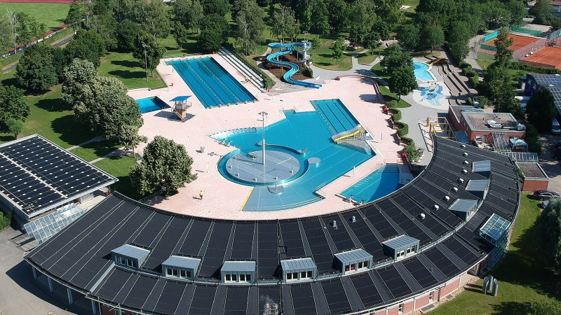 Luftaufnahme eines Freibads mit Becken, Rutschen und Solarzellen auf dem Dach. Umgeben von B&auml;umen und Sportpl&auml;tzen., &copy; Stadt Leonberg