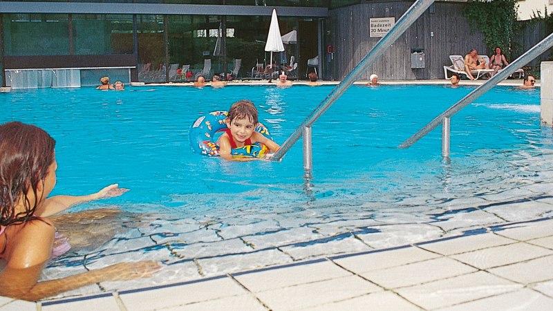 Ein Kind im Schwimmring im Pool, Erwachsene schwimmen und entspannen auf Liegestühlen im Badhaus Bad Boll., © Stuttgart-Marketing GmbH