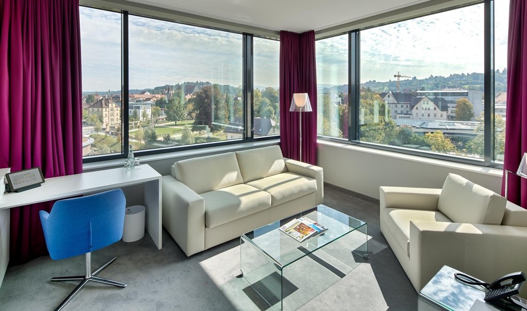 Moderne Suite mit großen Fenstern, weißen Sofas, Glastisch, Schreibtisch und lila Vorhängen. Blick auf eine Stadtlandschaft., © Hotel am Remspark Moderne Suite mit großen Fenstern, weißen Sofas, Glastisch, Schreibtisch und lila Vorhängen. Blick auf eine Stadtlandschaft., © Hotel am Remspark