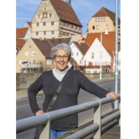 Eine ältere Frau steht lächelnd auf einer Brücke in Besigheim. Im Hintergrund sind malerische Fachwerkhäuser zu sehen., © Stadt Besigheim