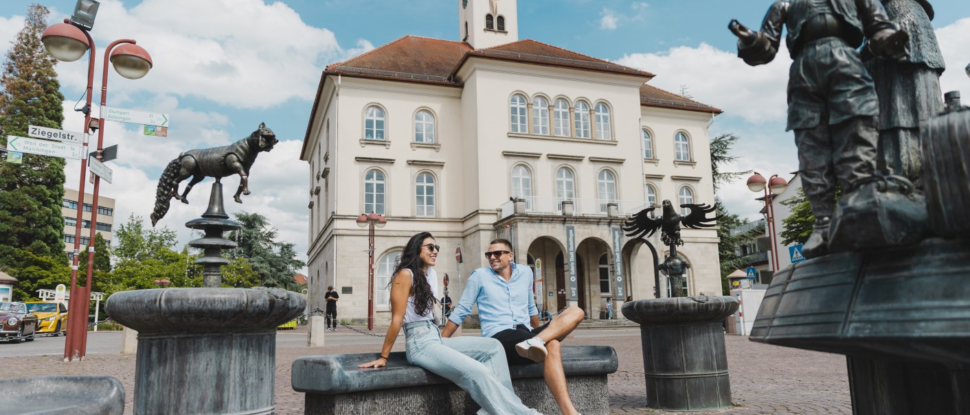 Zwei Personen sitzen vor einem historischen Gebäude in Sindelfingen. Im Vordergrund sind Skulpturen und ein Brunnen zu sehen. Der Himmel ist blau mit Wolken., © benjaminknoblauch.de Zwei Personen sitzen vor einem historischen Gebäude in Sindelfingen. Im Vordergrund sind Skulpturen und ein Brunnen zu sehen. Der Himmel ist blau mit Wolken., © benjaminknoblauch.de