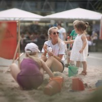 Eine Frau und zwei Kinder spielen im Sand an einem Stadtstrand. Die Frau isst Eis, während die Kinder mit Eimern und Schaufeln spielen., © Stadtmarketing Göppingen