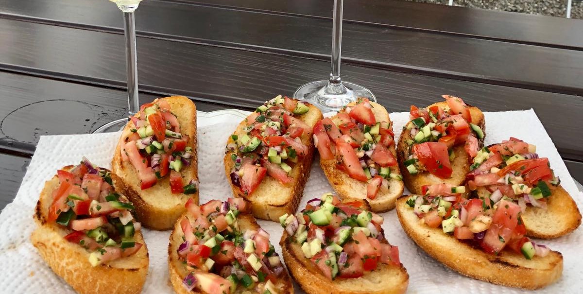 Zwei Gläser Wein, eines weiß und eines rot, stehen auf einem Tisch mit Bruschetta. Im Hintergrund sind Sonnenschirme und ein moderner Außenbereich zu sehen., © Salva's Curry-Truck