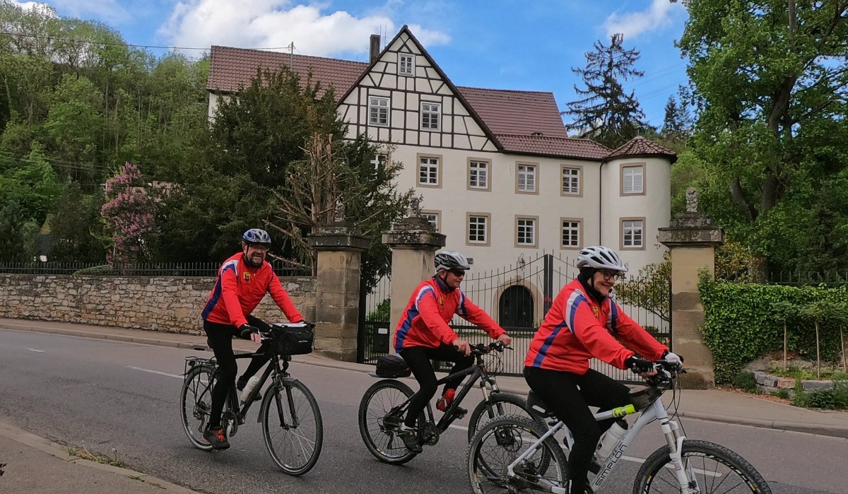 Drei Radfahrer in roten Jacken fahren auf einer Straße vor einem großen Fachwerkhaus mit Garten., © Land der 1000 Hügel - Kraichgau-Stromberg Drei Radfahrer in roten Jacken fahren auf einer Straße vor einem großen Fachwerkhaus mit Garten., © Land der 1000 Hügel - Kraichgau-Stromberg