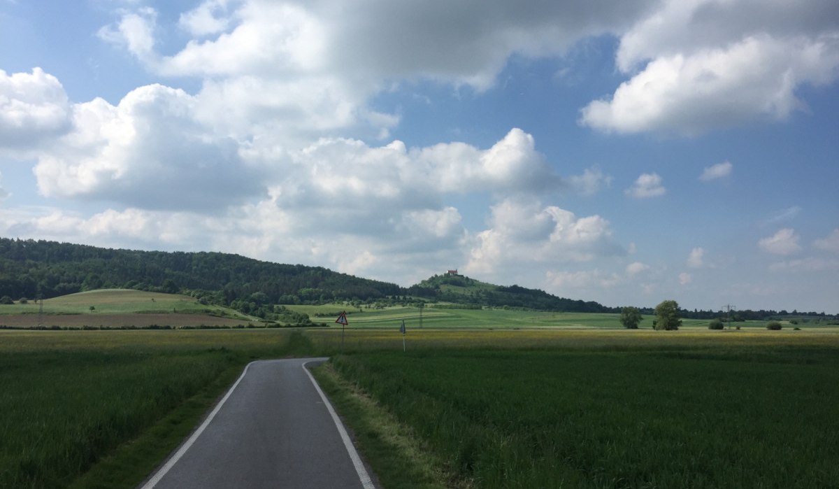 Ein Radweg schlängelt sich durch grüne Felder, im Hintergrund die Wurmlinger Kapelle auf einem Hügel unter einem bewölkten Himmel., © www.pro-cycl.de Ein Radweg schlängelt sich durch grüne Felder, im Hintergrund die Wurmlinger Kapelle auf einem Hügel unter einem bewölkten Himmel., © www.pro-cycl.de