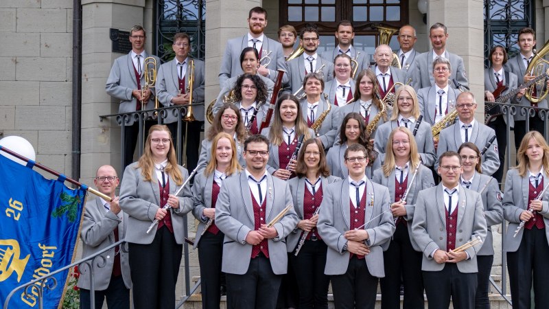 Gruppenfoto der Musiker der Stadtkapelle mit dunkelroter Weste, grauem Jacket und schwarzen Hosen. Die Musiker stehen auf den Treppenstufen vor dem Eingang des Neuen Schlosses und halten Musikinsturmente in den Händen, © Stadtkapelle Gaildorf