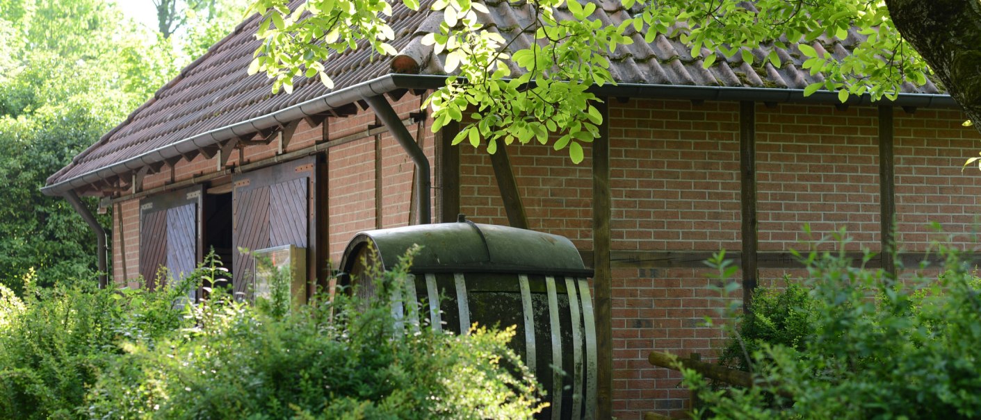 Backsteingeb&auml;ude mit Holzt&uuml;ren, umgeben von gr&uuml;nen B&uuml;schen und B&auml;umen. Ein altes Fass steht im Vordergrund., &copy; Frank Nonnenmann