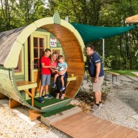 Eine Familie sitzt vor einem fassförmigen Holzhaus im Freien. Ein Mann steht daneben. Umgeben von Bäumen und einem Holzweg., © Remsfässer