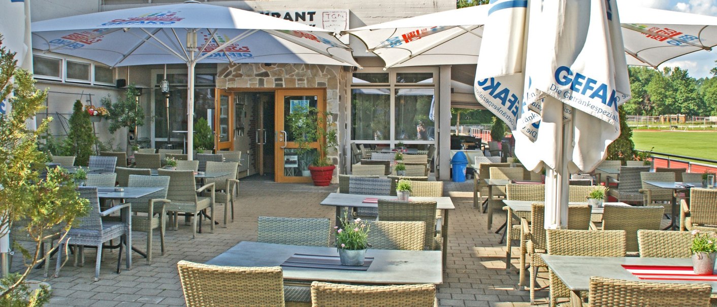 Gemütlicher Außenbereich eines Restaurants mit geflochtenen Stühlen, Tischen und großen Sonnenschirmen. Im Hintergrund ein Gebäude mit roter Fassade., © Restaurant Neckarau, P. Kotaidis