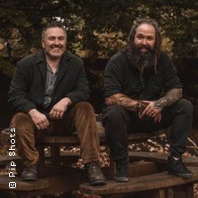 Zwei M&auml;nner sitzen l&auml;chelnd auf einer Holzbank im Freien, umgeben von herbstlichen B&auml;umen. Beide tragen l&auml;ssige Kleidung., &copy; links im Bild