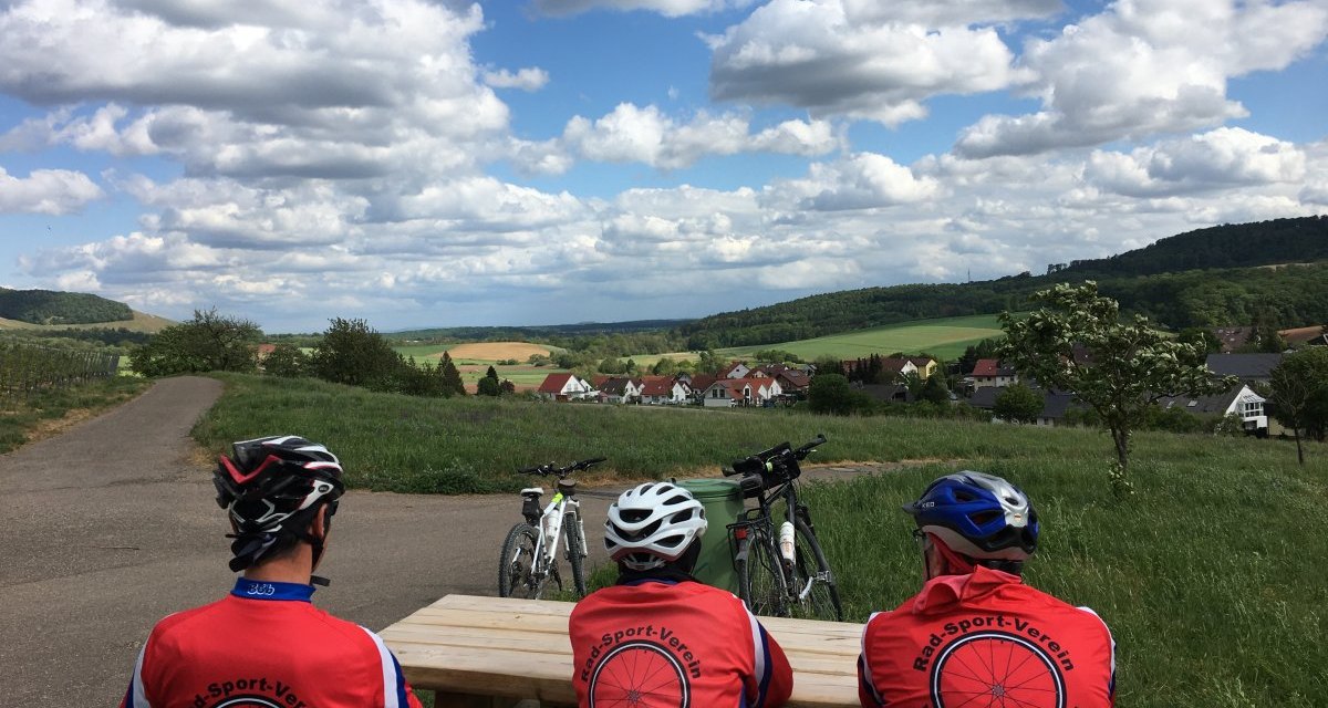 Drei Radfahrer in roten Trikots sitzen auf einer Bank und blicken auf eine ländliche Landschaft mit Feldern und Hügeln unter einem bewölkten Himmel., © Land der 1000 Hügel - Kraichgau-Stromberg Drei Radfahrer in roten Trikots sitzen auf einer Bank und blicken auf eine ländliche Landschaft mit Feldern und Hügeln unter einem bewölkten Himmel., © Land der 1000 Hügel - Kraichgau-Stromberg