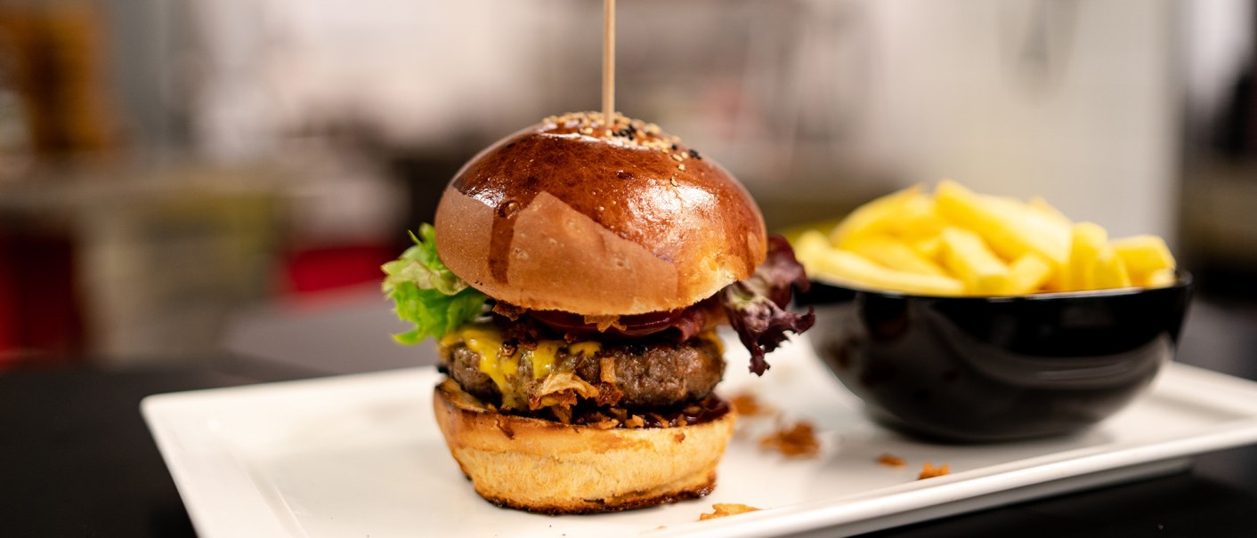 Ein saftiger Burger mit Salat und Käse auf einem Teller, daneben eine Schale mit Pommes frites., © Sensapolis GmbH
