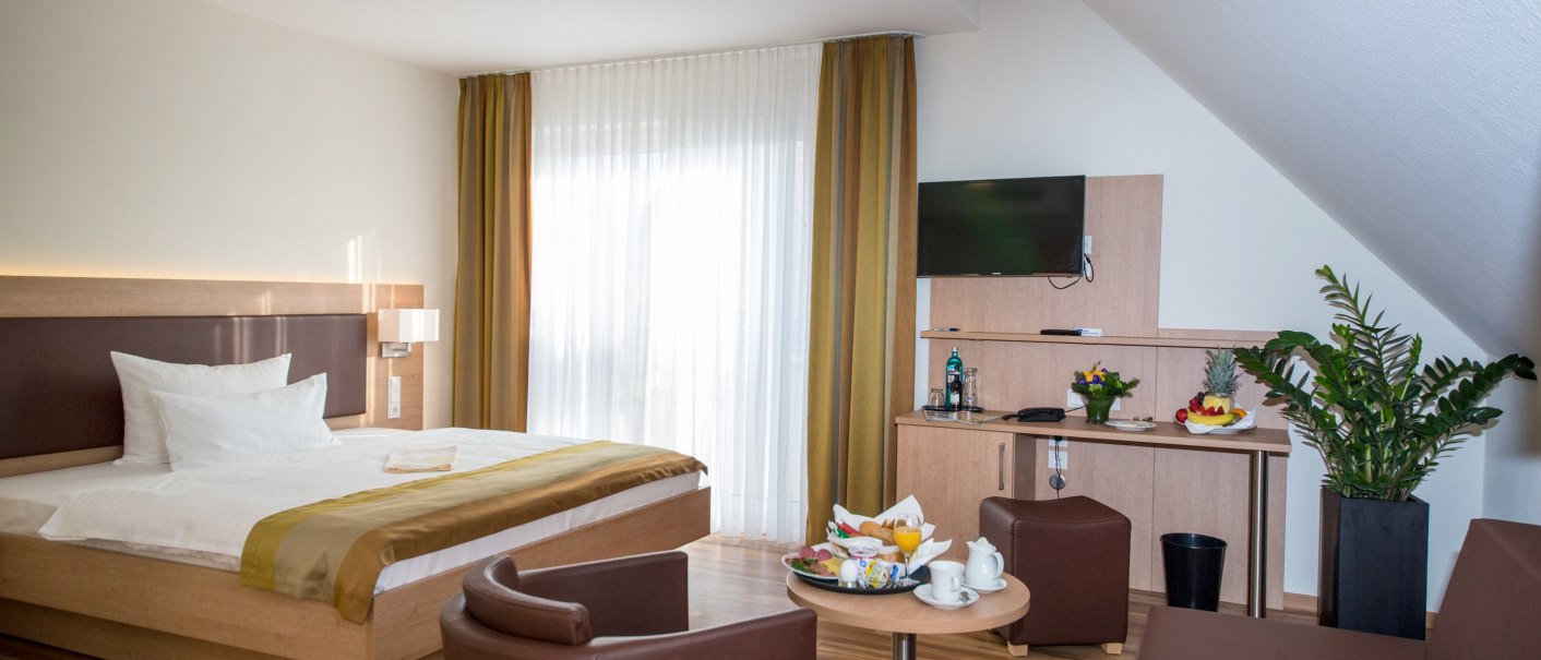 Modernes Doppelzimmer mit großem Bett, Tisch, zwei Sesseln, TV und Pflanzen. Helle Vorhänge und Holzmöbel schaffen eine gemütliche Atmosphäre., © benjamin stollenberg | fotografie Modernes Doppelzimmer mit großem Bett, Tisch, zwei Sesseln, TV und Pflanzen. Helle Vorhänge und Holzmöbel schaffen eine gemütliche Atmosphäre., © benjamin stollenberg | fotografie