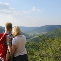 Ein Paar mit rotem Rucksack genießt den Ausblick vom Eppenzillfelsen auf eine grüne Hügellandschaft unter blauem Himmel., © Bad urach Tourismus